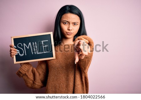 Young chinese woman holding blackboard with smile word over isolated pink background pointing with finger to the camera and to you, hand sign, positive and confident gesture from the front