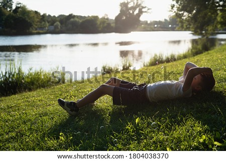 boy lies on  green grass, rest by  river,  sunset yellow light,