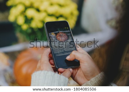 Young woman taking photo of pumpkins and autumn flowers. Girl photographing on phone rustic halloween street decor, hands close up. Happy Thanksgiving and Halloween