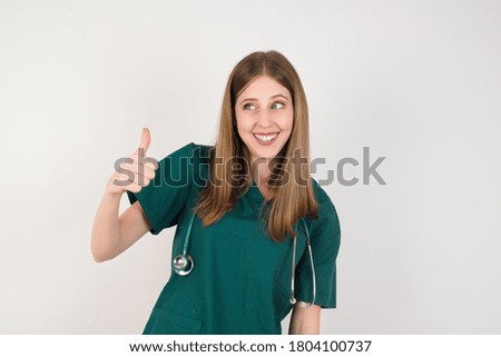Female doctor wearing a green scrubs and stethoscope is on white background Looking proud, smiling doing thumbs up gesture to the side. Good job!