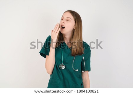 Female doctor wearing a green scrubs and stethoscope is on white background being tired and yawning after spending all day at work.