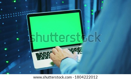 IT technician is working on laptop at row of server racks, watching template green monitor and running diagnostics, staying at futuristic server center. Database technology. Royalty-Free Stock Photo #1804222012