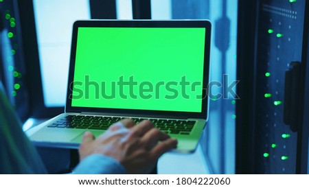 IT administrator using a mock-up laptop running diagnostics and web hosting with chrome key greenscreen device inside server cabinet of database. Close-up. Advanced data center. Royalty-Free Stock Photo #1804222060