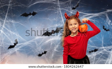 halloween, holiday and childhood concept - smiling girl in party costume with red devil's horns holding jack-o-lantern pumpkin over bats and cobweb in night sky on background