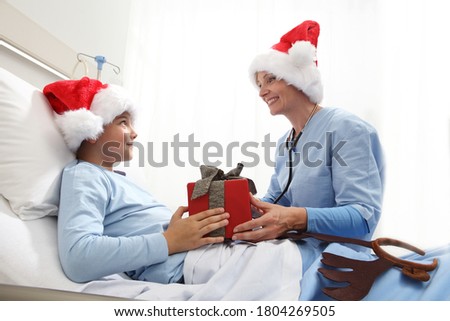 Christmas holiday in hospital happy child lying in bed with Santa Claus hat and nurse giving him a present 