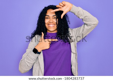 Young african american sporty woman wearing casual sweatshirt over purple background smiling making frame with hands and fingers with happy face. Creativity and photography concept.