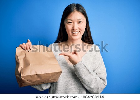 Young asian woman holding delivery paper bag for takeaway food over blue isolated background very happy pointing with hand and finger