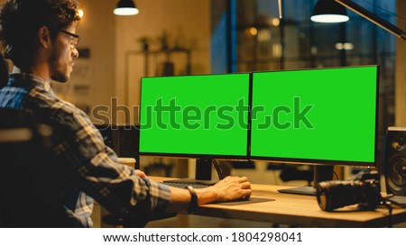 Over the Shoulder: Creative Designer Sitting at His Desk Uses Desktop Computer with Two Green Mock-up Screens. Professional Office Employee Working Late in the Evening in His Studio