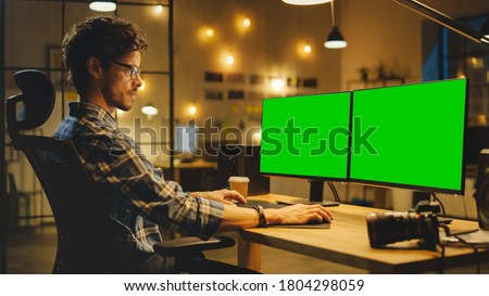 Late Evening in Creative Office: Professional Photographer Works on a Desktop Computer with Two Green Mock-up Screens. Modern Studio Office with Hanging Lightbulbs