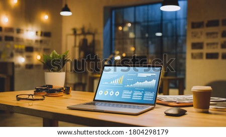 Laptop on the Desk in the Office Shows Company Growth Data with Graphs, Charts, Software UI. In the Background Stylish Modern Office Studio in the Evening with Big Cityscape Window