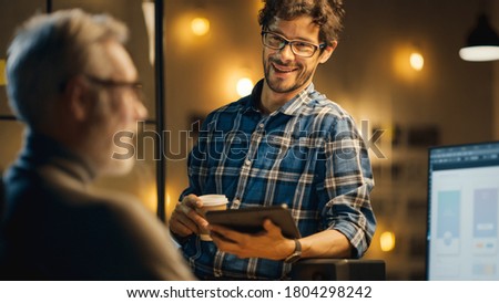 In the Evening Creative Middle Aged Smartphone Software Developer Works on a Desktop Computer with Screen Showing Application Design, Has Conversation with Younger Specialist who Uses Tablet Computer