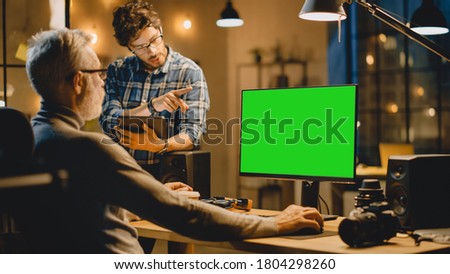 In the Evening Creative Middle Aged Man Works on a Desktop Computer Green Mock-up Screen, Has Conversation with Younger Specialist who Uses Tablet Computer. Stylish Big City Office late in the Evening