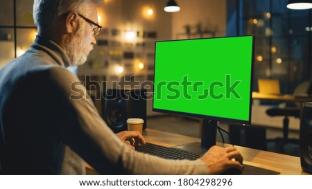 Creative Middle Aged Designer Sitting at His Desk Uses Desktop Computer with Green Mock-up Screen. In the Evening Creative Employe Working on Computers in the Office with Cityscape Window View