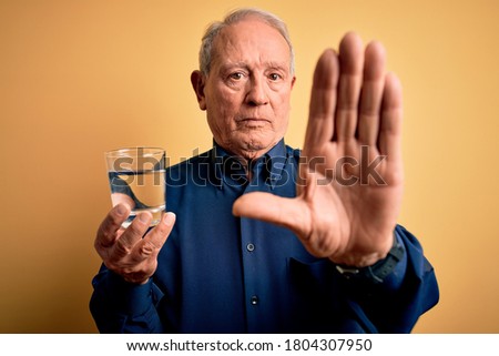 Grey haired senior man drinking a fresh glass of water over yellow isolated background with open hand doing stop sign with serious and confident expression, defense gesture