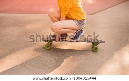 Unrecognizable teen skater sitting on haunches on skateboard while spending weekend day in skate park
