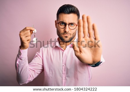Young real estate business man holding new house keys over pink background with open hand doing stop sign with serious and confident expression, defense gesture