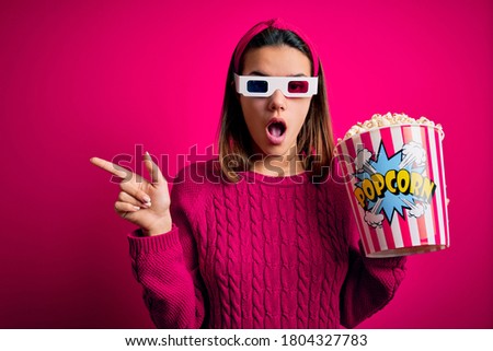 Young beautiful girl watching movie using 3d glasses eating box with popcorns Surprised pointing with finger to the side, open mouth amazed expression.