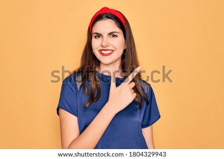 Young beautiful brunette woman wearing red lips over yellow background cheerful with a smile of face pointing with hand and finger up to the side with happy and natural expression on face