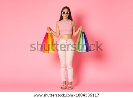 Full length portrait of stylish, pretty asian woman travelling abroad and buying souvenirs, new clothes in stores, carry shopping bags, wearing sunglasses and smiling over pink background