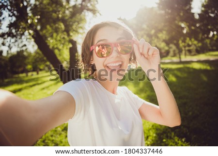 Closeup headshot photo of careless charming pretty lady shiny toothy smiling hold camera spectacles take selfie open mouth send picture friend abroad park wear sunglass white shirt outdoors