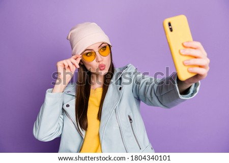 Photo of funny pretty youngster lady hold telephone make selfies popular blogger sending air kisses wear sun specs casual hat jacket yellow pullover isolated purple color background