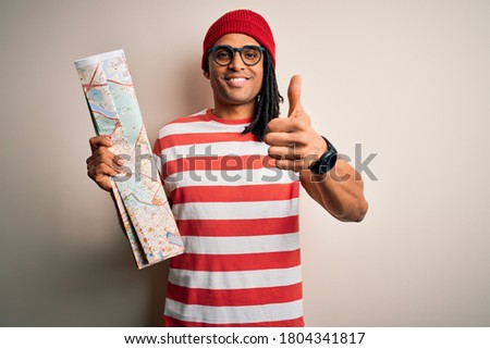 Young african american afro tourist man with dreadlocks wearing glasses holding city map happy with big smile doing ok sign, thumb up with fingers, excellent sign