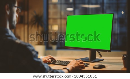 Over the Shoulder: Creative Young Man Sitting at His Desk Using Desktop Computer with Mock-up Green Screen. Evening in the Stylish Office Studio with City Window View Royalty-Free Stock Photo #1804351051
