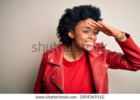 Young beautiful African American afro woman with curly hair wearing casual red jacket very happy and smiling looking far away with hand over head. Searching concept.