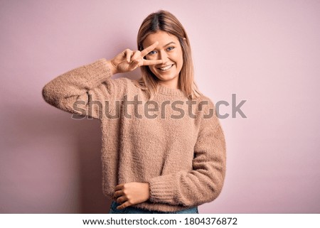 Young beautiful blonde woman wearing winter wool sweater over pink isolated background Doing peace symbol with fingers over face, smiling cheerful showing victory