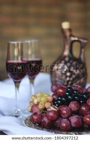 A tray with bunches of grapes of different varieties on which are wedding rings. Against the Background of a Greek-style Bottle and two glasses of red wine. Blur soft focus