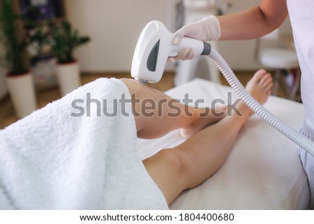 Beautician doing epilation on beautiful girl's leg in medical center. Female receiving laser light hair removal treatment for hairless smooth skin at cosmetology salon. Close up Royalty-Free Stock Photo #1804400680