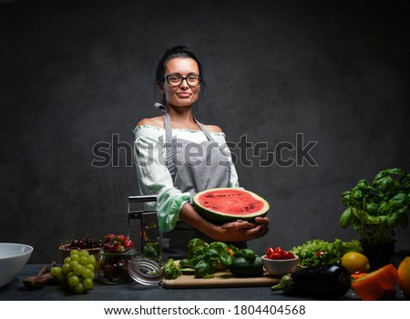 Middle-aged female chef holds half the watermelon in kitchen, smiling and looking on the camera. Healthy and proper nutrition on a diet. Studio photo on a dark background