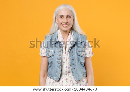 Smiling cheerful beautiful elderly gray-haired woman 70s with long hair wearing casual dress denim waistcoat looking camera isolated on yellow color background, studio portrait.