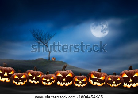 Lots of halloween lanterns with evil face and eyes, Jack O Lantern, against a spooky looking landscape background at night with a glowing full moon, lone tree and cloudy sky.