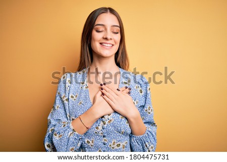 Young beautiful brunette woman wearing casual floral t-shirt standing over yellow background smiling with hands on chest with closed eyes and grateful gesture on face. Health concept.