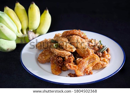 Fried banana mix sesame and pandan leaf for aroma (Thai called Kluay Thod) in white dish and Banana on black cloth background Thai snack / Select focused space for text