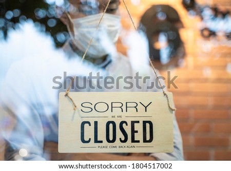 Waiter in uniform wearing a white mask is standing at entrance door of a restaurant. The door hangs a wooden sign "sorry we are closed". Lock down business  during covid19 crisis is new normal.