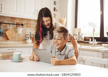 Happy teenagers brother and sister are sitting at a table in the kitchen with a tablet on the network, chatting with friends, watching a cartoon. Modern child