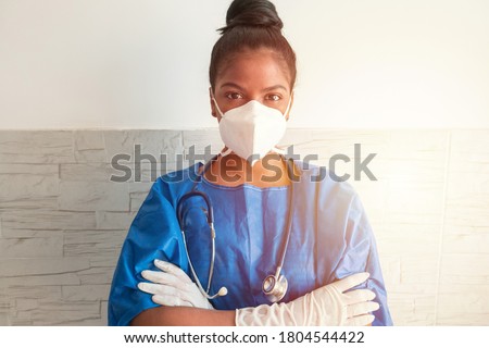 A young African nurse wears a blue uniform, a mask and gloves while standing with her arms crossed in the hospital.