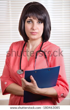 Beautiful young woman doctor holding a clipboard with patient records pausing on her ward rounds to look at the camera with a serious expression
