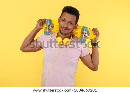 Hesitating african american young man wearing headphones, holding skateboard on shoulders over yellow background.