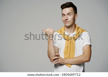 a man of Caucasian appearance, shows a friend interesting things, makes a finger gesture to the side. stylish brunette hairstyle. white t-shirt and yellow jacket on the shoulders. grey background phot