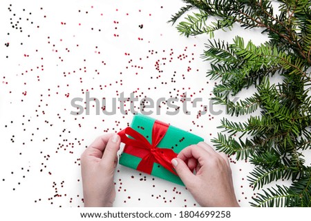 Hands hold one Christmas gift with red ribbon on the white table with Christmas tree nearby . Christmas wrapping