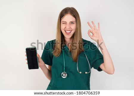 Portrait Female doctor wearing a green scrubs and stethoscope is on white background holding in hands cell showing ok-sign  Royalty-Free Stock Photo #1804706359