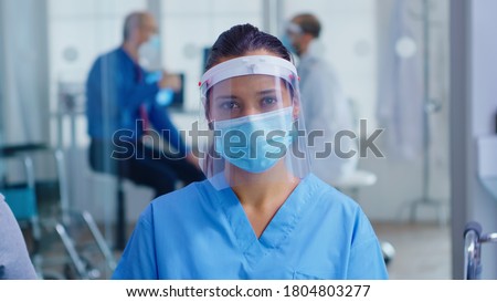 Medical assistant with visor and face mask against coronavirus looking at camera in hospital waiting area. Doctor consulting senior man in examination room. Royalty-Free Stock Photo #1804803277