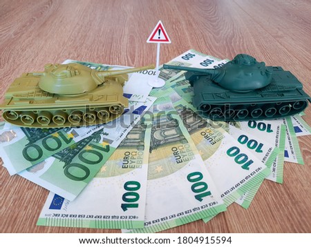 Two model plastic toy tanks and warning traffic sign on a pile of euro banknotes against wooden background