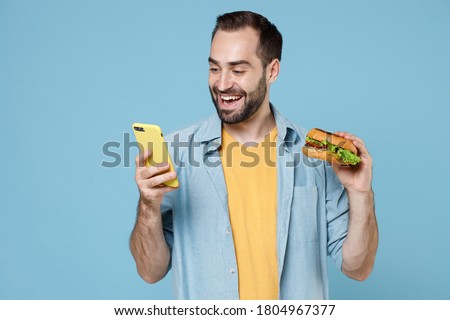 Smiling cheerful young bearded man 20s in casual clothes holding in hands american classic fast food burger and using mobile cell phone, typing sms isolated on pastel blue background, studio portrait.