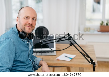 Middle-aged man recording a podcast seated at a desk turning to smile at the computer with his headphones around his neck with copy space