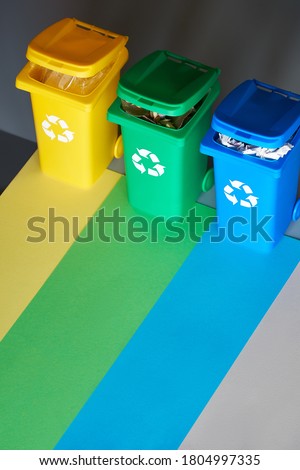 Three color coded recycle bins, isometric picture on geometric layered paper background. Recycling sign on blue, yellow and green containers. Waste separation, reduce mixed waste and recycle plastic.
