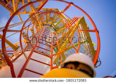 Low angle view of an engineer climbing a tower on an industrial steel ladder with safety cage. Heavy industry place of work with yellow, orange and red steel colors as safety first concept. Royalty-Free Stock Photo #1805172085